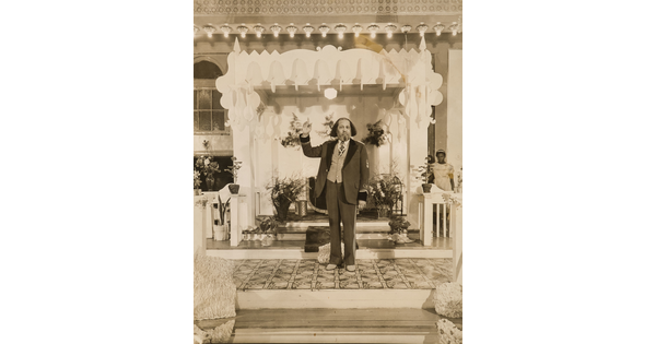 A sepia-toned portrait photograph of a Black man with mid-length coiled hair, in a three-piece suit with his arm raised and index finger pointing up, and an altar with flower arrangements behind him.