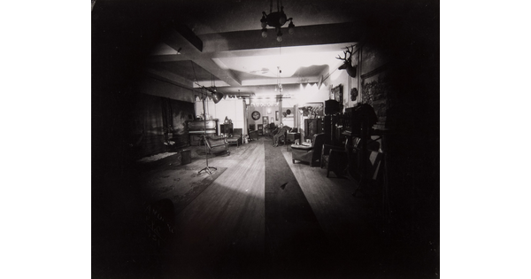 A dark black-and-white photograph of a living room with a spotlight on a man seated in an armchair in a far corner.