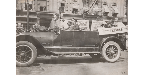 A black-and-white photograph of a convertible vehicle on an urban street with four Black people in the car, a banner draped on the side, and signs displayed in the background.