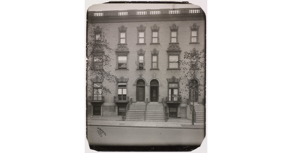 A black-and-white photograph of the facades of three-story row homes with steps leading to front doors.