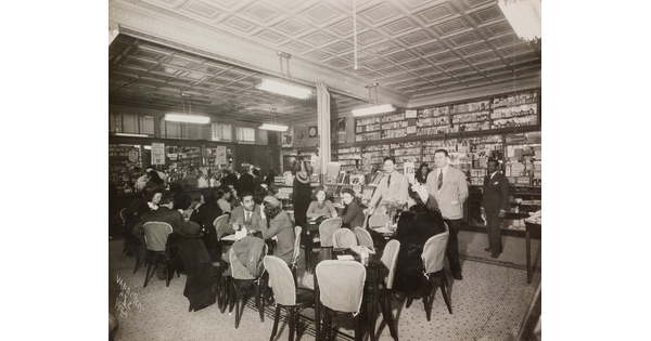 A black-and-white photograph of the interior of a soda fountain filled with a crowd of men and woman socializing.