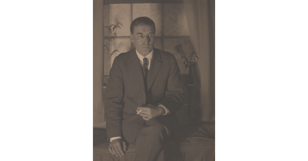 A sepia-toned photograph of a White man in a suit, legs crossed, holding a cigarette in one hand that is resting on his leg, seated in front of a window.