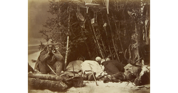 A black-and-white photograph of several men asleep outside at a winter campsite, with their snow gear stored in branches of trees.