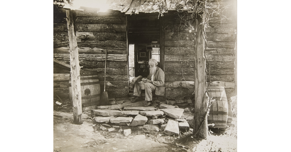 A black-and-white photograph of an old White man with a long beard sitting in the doorway of a log cabin.