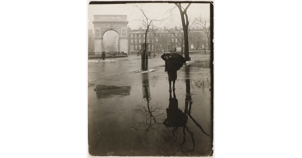 A black-and-white photograph of someone walking on wet pavement through a plaza with bare trees and a white arch in the distance.