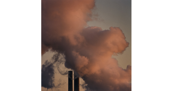 A color photograph of pink smoke against a gray sky above two industrial smokestacks.