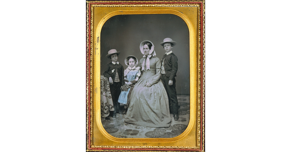 A hand-painted black-and-white formal portrait photograph in a gold frame of a White woman sitting with two boys and a girl standing around her.