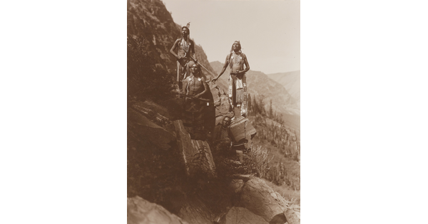A sepia-toned photograph of four Native Americans in traditional dress arranged on a steep, rocky slope.