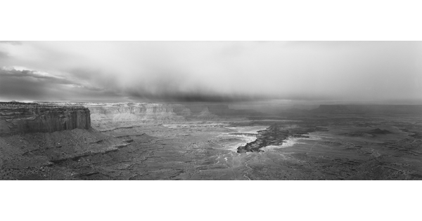 A black-and-white panoramic photograph of a steep canyon under a stormy sky.