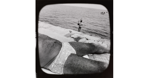 A black-and-white photograph of the hairy torso of a man wearing a brief-style swimsuit sunbathing near a cement ledge by water where a fully-clothed person plays bagpipes in the background.