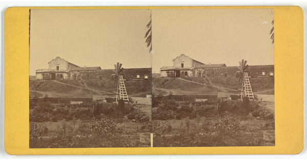 Two almost identical side-by-side sepia-toned photographs of an adobe building on a hill with a windmill in the foreground.