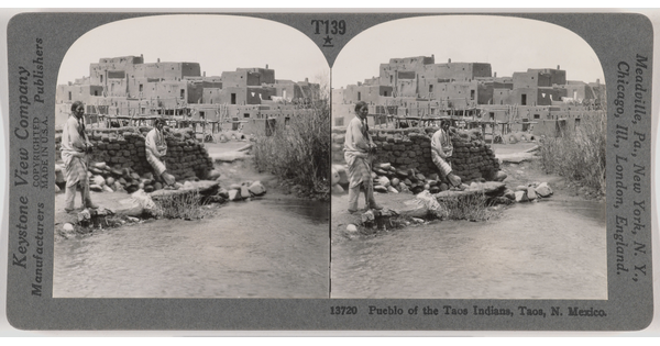 Two almost identical side-by-side black-and-white photographs of two Native Americans standing in front of a pile of mud bricks with an adobe pueblo in the background.