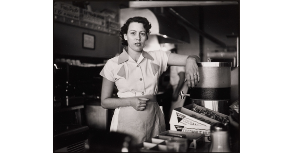 A black-and-white photograph of a White woman in a waitress uniform standing behind a diner counter with an arm propped on drink dispenser.