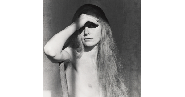 A black-and-white portrait photograph of a nude White woman with long hair using her hand to shield her eyes.