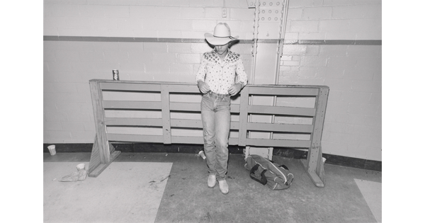 A black-and-white photograph of a teenager wearing a cowboy hat and a decorative western-style shirt leaning back against a showjumping fence.
