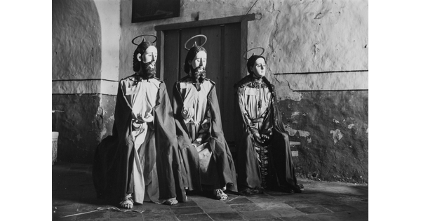 A black-and-white photograph of three seated mannequins, two with beards, wearing tunics and loose robes, sandals, and with halos mounted on their heads.