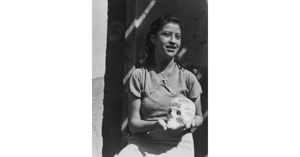 A black-and-white photograph of a brown-skinned girl showing the decorated sugar skull she holds in her hands.