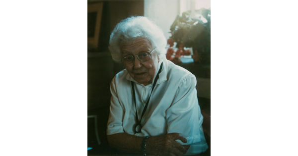 A color photograph of an older White woman with white hair, glasses, and a white blouse leaning forward with her arms crossed.