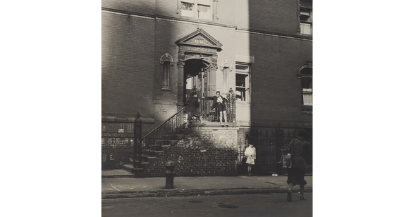 A black-and-white photograph of the entrance to an apartment building with children playing on the outside of the railing on the steps that lead to the door.