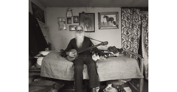 A black-and-white photograph of an elderly White man with a long white beard sitting on a bed in a cozy bedroom holding a banjo with one hand, the other hand around a cat lying next to him.