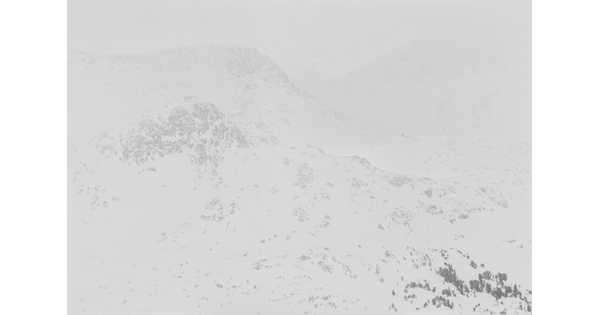 A black-and-white photograph of a snowy mountain with snow and clouds slightly obscuring the view.