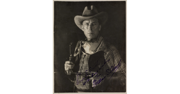 A black-and-white photograph of a White man in western attire holding a pistol in each hand, an autograph in blue in across the bottom.