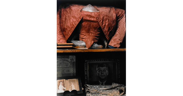 A color photograph of a shelf displaying a red shirt and jacket, photographs of two women, a portrait of John F. Kennedy, some folded fabric, and a Boy Scouts of America certificate.
