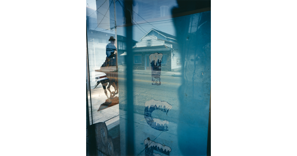 A color photograph taken through a window by the side of an ice machine and a poster of a man on a horse, as well as reflections of a house and other buildings.