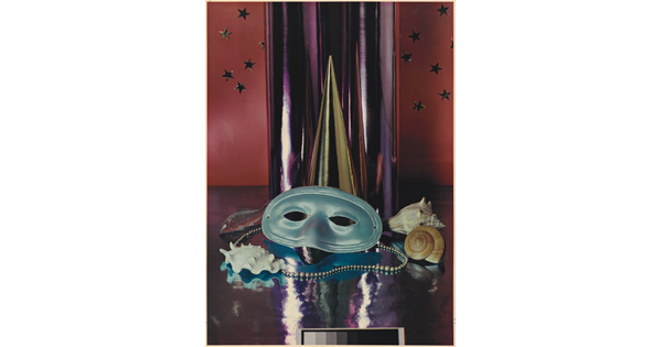 A color photograph of a simple party mask, Mardi Gras beads, and seashells in front of a reflective silver cylinder.