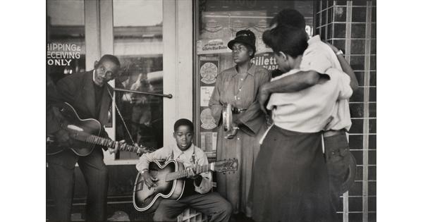 A black-and-white photograph of Black adults and boy playing instruments in front of a storefront while a Black couple watches.