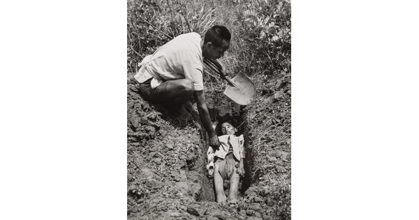 A black-and-white photograph of an Asian man holding a shovel crouched down next to a freshly dug grave containing a emaciated body.