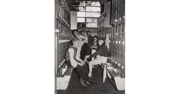 A black-and-white photograph of several men in various states of dress in the row of a locker room.