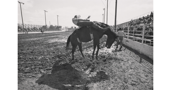 A black-and-white photograph of a person riding bareback on a rearing horse in a dirt arena.