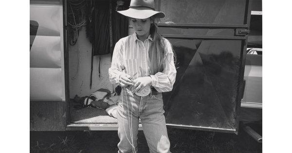 A black-and-white photograph of a young White woman with long hair wearing western clothing cutting rope with a knife.