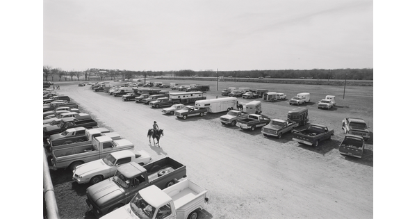 A black-and-white photograph of a single rider on horseback amid rows of parked cars and trucks.