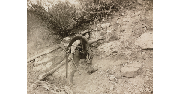 A black-and-white photograph of a White man climbing a rocky ridge holding a rifle with a mountain lion carcass over his shoulder.