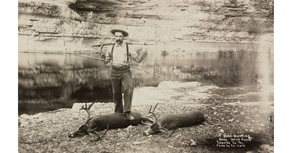 A black-and-white photograph of a White man racking a rifle across his shoulders while standing over two buck carcasses next to a creek.