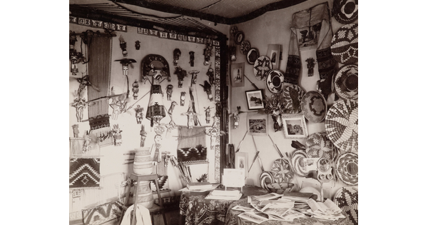 A black-and-white photograph of an interior room, the walls covered with Native American dolls, baskets, blankets, and pottery.