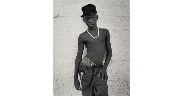 A black-and-white portrait photograph of a Black teenage boy wearing a backward baseball cap, a necklace, no shirt, and loose fitting jeans against a concrete wall.