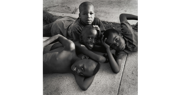A black-and-white photograph of four young Black boys lying on each other and a sidewalk, smiling up at the viewer.