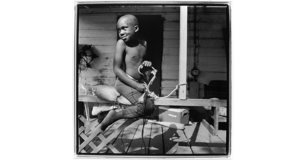 A black-and-white photograph of a young Black boy straddling the railing of a porch pretending to ride a horse.