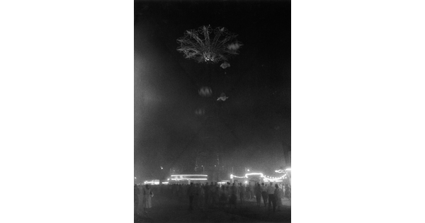 A black-and-white photograph looking up at a tall ride on festival grounds at night.