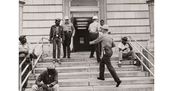 A black-and-white photograph of Black men on steps, two holding protest signs, and two White police officers approaching them, one with a baton.