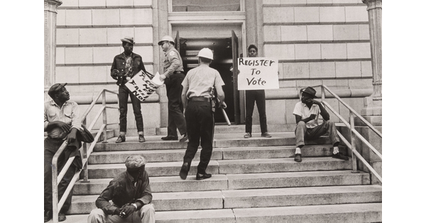 A black-and-white photograph of Black men on steps, two holding protest signs, and two White police officers approaching them, one with a baton.