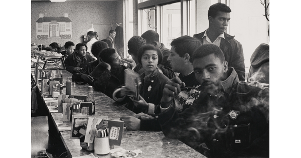 A black-and-white photograph of mostly young Black people and one White person at a crowded diner counter, talking and smoking.