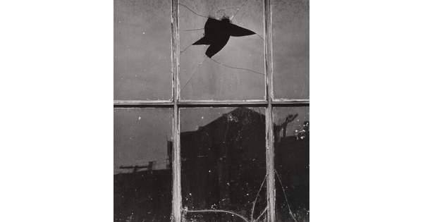 A black-and-white photograph of dirty and cracked windowpanes, one with a hole through it, and a silhouette of a building reflected in the glass.