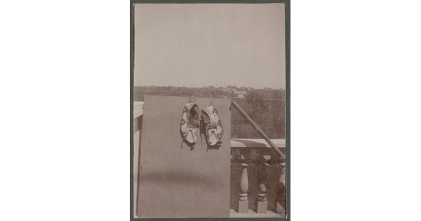 A black-and-white photograph of a pair of beaded moccasins hanging on a board that is leaning on a balustrade; tree-covered hills in the distance.