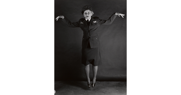 A black-and-white portrait photograph, in full length, of an older White woman wearing military uniform, jumping and striking a flying-pose with her arms.