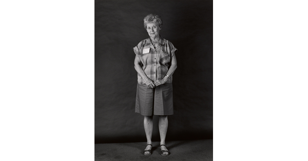 A black-and-white portrait photograph, in full length, of an older White woman with a shy smile wearing a short-sleeved plaid shirt and culotte shorts.