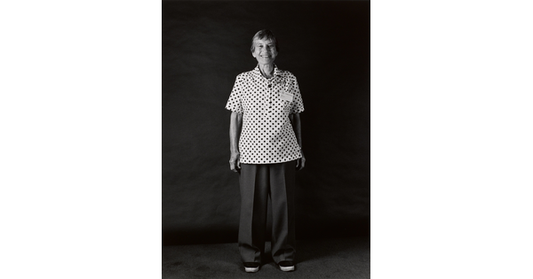 A black-and-white portrait photograph, in full length, of a smiling older White woman wearing  a short-sleeved polka dot shirt and long pants.